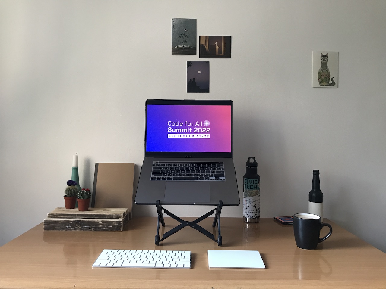 A work space showing a desk, coffee cup, water bottle, illustrations on the wall, and a laptop displaying the Code for All Summit poster